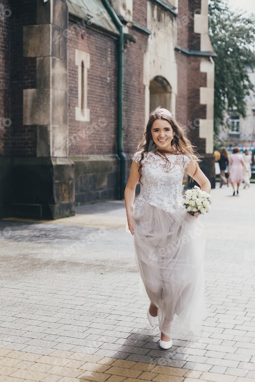 Preview: Beautiful happy bride in stylish vintage dress running in sunny street on background of church