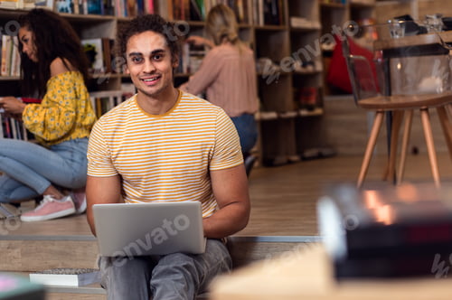 Preview: Portrait of young male student sitting at library using laptop with his friends in background.
