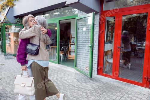 Preview: Senior lady hugs smiling friend with glasses meeting on modern city street