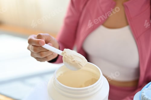 Preview: Young woman in sportswear hand holding scoop making protein shake after morning workout.