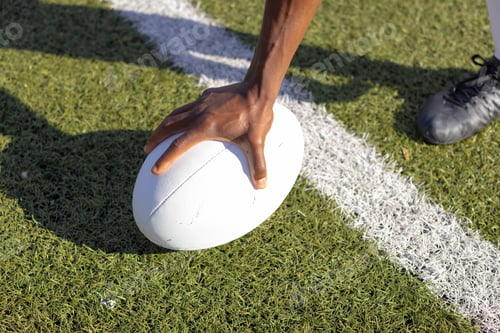 Preview: An African American young male athlete wearing cleats placing hand on rugby ball on grass field