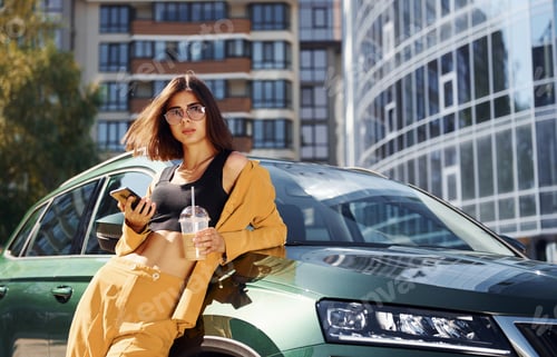 Preview: With smartphone. Young fashionable woman in burgundy colored coat at daytime with her car