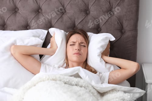 Preview: Young woman covering ears with pillow while trying to sleep in bed at home. Early morning