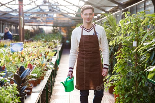 Preview: Smiling Man Gardener In Glasses And Brown Apron Standing And Holding Watering Can In Orangery