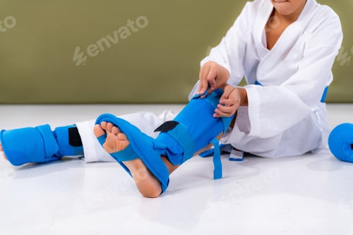Preview: Boy wearing blue protective gear putting guards on legs before karate sparring match.