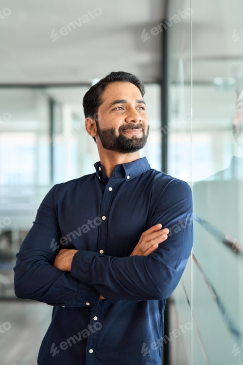 Preview: Smiling Indian business man leader looking away standing in office hallway.