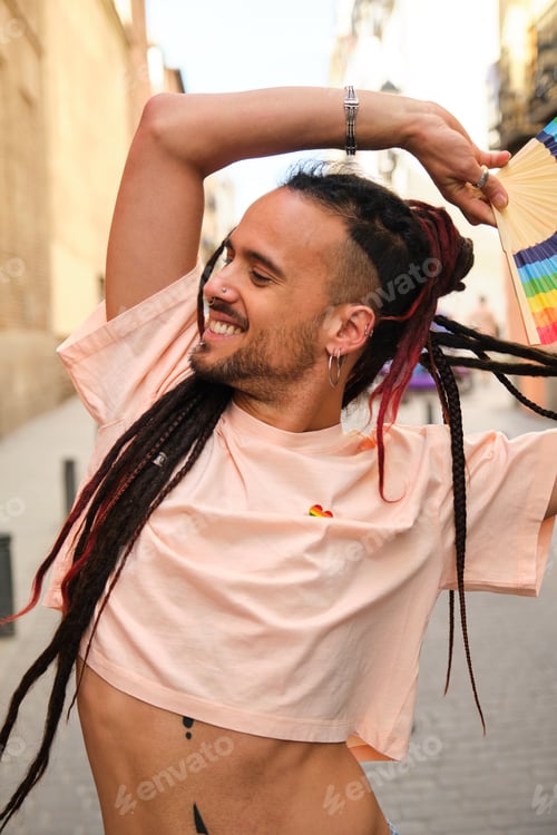 Preview: Gay man with dreadlocks smiling and dancing holding a rainbow fan.