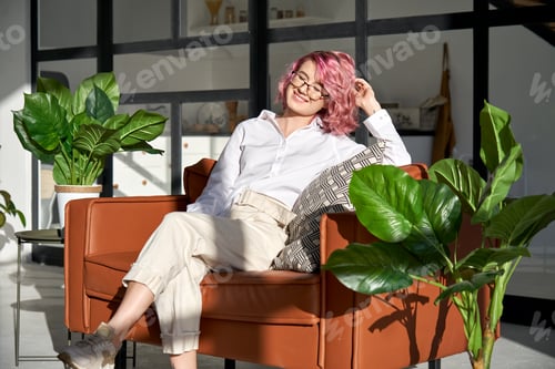 Preview: Smiling hipster woman with pink hair relaxing on sofa in sunny cozy living room.