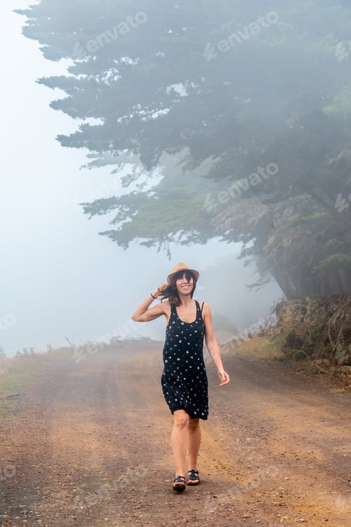 Preview: Tourist woman with hat walking through the foggy path towards the juniper forest El Hierro. Canary