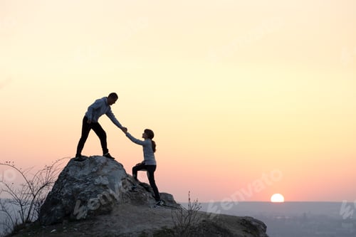 Preview: Man and woman hikers helping each other to climb stone at sunset in mountains. Couple climbing on