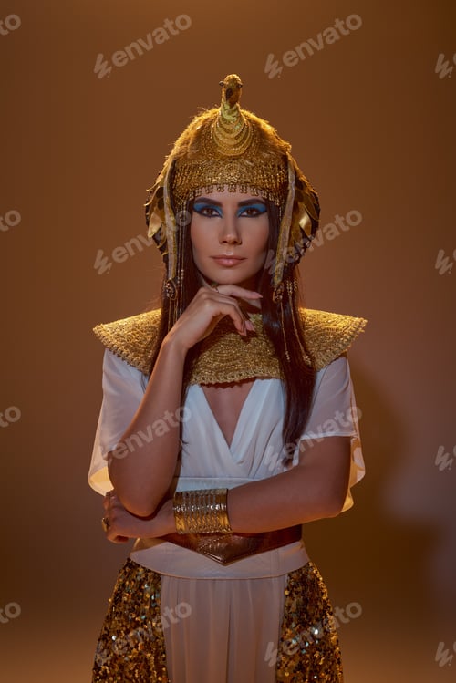Preview: Stylish brunette woman with makeup and egyptian attire looking at camera on brown background