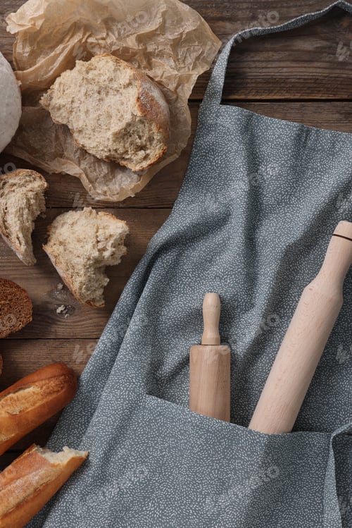 Preview: Clean kitchen apron with rolling pins and different types of bread on wooden table, flat lay