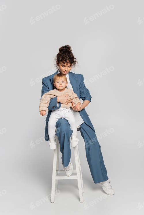 Preview: curly businesswoman in blue suit sitting with toddler daughter on high chair on grey