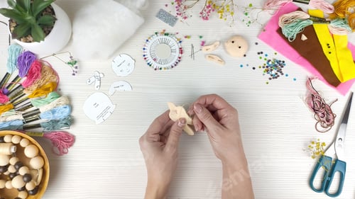 Preview: Female hands sew a handmade felt doll on white wooden table background.