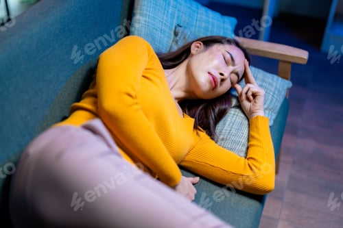 Preview: Asian woman resting on sofa, pressing hand against forehead, experiencing intense migraine pain