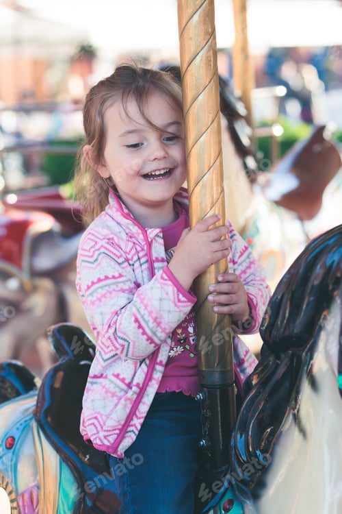 Preview: Little adorable smiling girl riding a horse on roundabout carousel at funfair