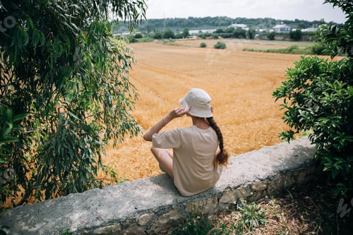 Preview: a teenage girl with a braid in a t-short and hat sits outdoor and looks at the wheat field