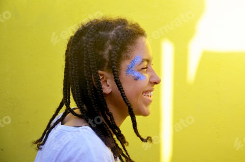 Preview: Teenage mixed race girl with blue glitter on her face , laughing, yellow background
