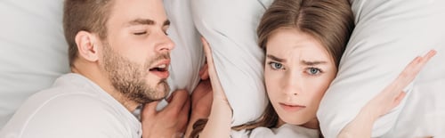 Preview: top view of exhausted woman plugging ears with pillow while lying in bed near snoring husband,