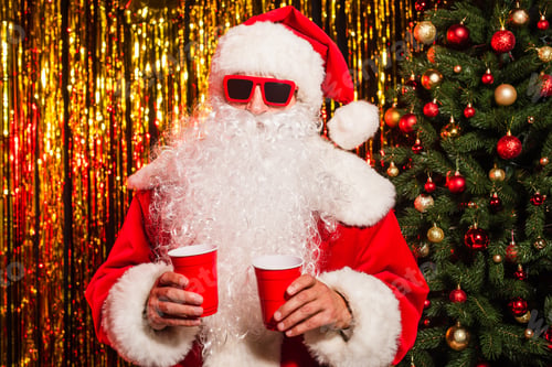 Preview: Santa claus in sunglasses and hat holding plastic cups near christmas tree and tinsel