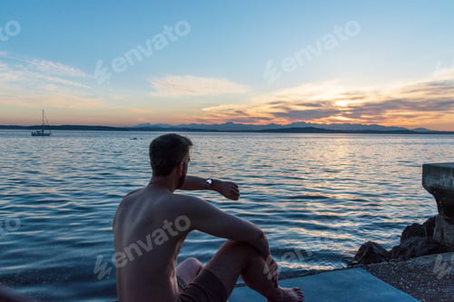 Preview: Man sitting on yoga mat on sea wall at sunset checking smart watch sun dial.