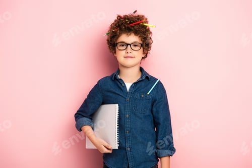 Preview: pleased kid in glasses with color pencils in curly hair holding notepad on pink