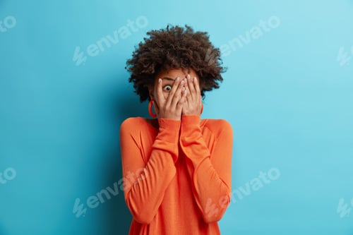 Preview: Scared African American woman covers face stares through fingers afraids of looking at something ter
