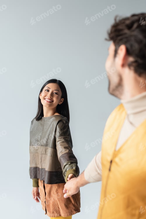 Preview: cheerful asian woman in leather pullover holding hands with blurred man isolated on grey