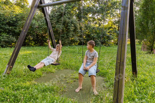 Preview: Two kids are playing joyfully on a swing set in a lush green area filled with trees