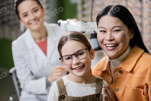 Preview: happy kid in eyeglasses looking at camera near asian mom and blurred ophthalmologist in optics shop