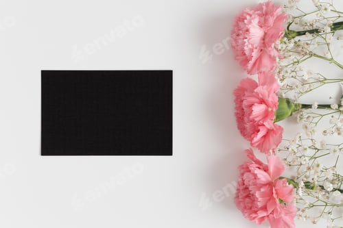 Preview: Top view of a black card mockup with gypsophila and pink carnations on a white table.