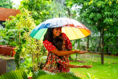 Preview: Two Indian girls playfully enjoy monsoon rain under umbrella, sharing joyful outdoor moment