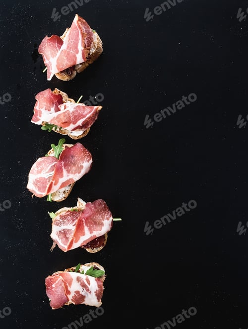 Preview: Bruschettas with prosciutto smoked meat, dried tomatoes and arugula over black backdrop, top view
