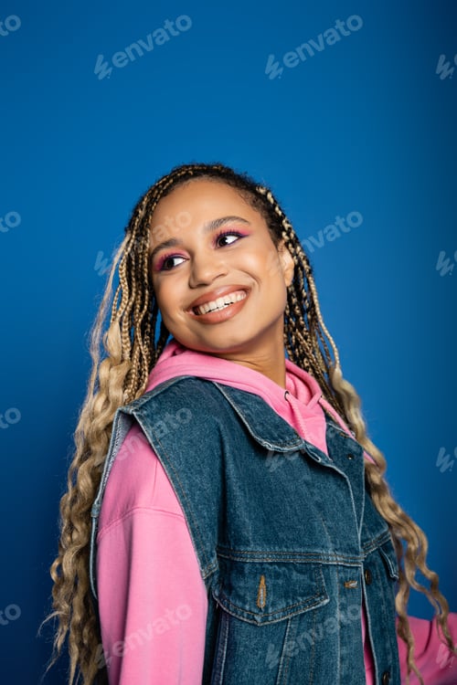 Preview: happy african american woman with dreadlocks and pink hoodie smiling and looking away on blue