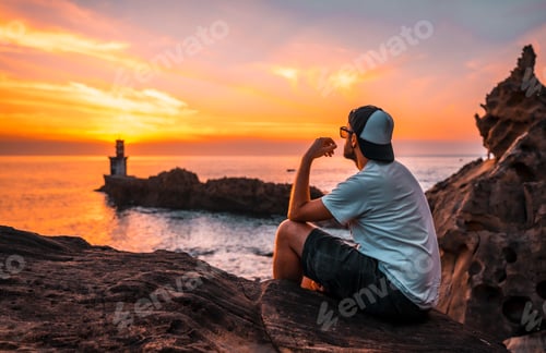 Preview: A young man sitting in a white shirt and cap looking at the orange sunset
