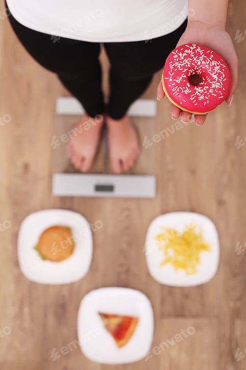 Preview: Young beautiful woman standing on scales and holding a donut.