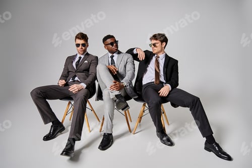 Preview: appealing interracial men with sunglasses in smart attires sitting on chairs on gray backdrop