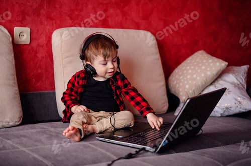 Preview: Little toddler boy sits on sofa wathes cartoons on laptop using headset