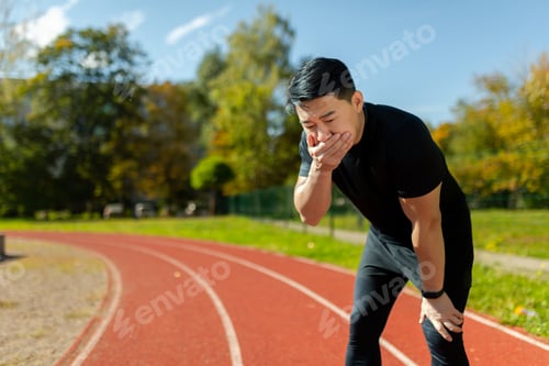 Preview: Asian young man doing sports at the stadium. He stands bent over on the treadmill, covers his mouth