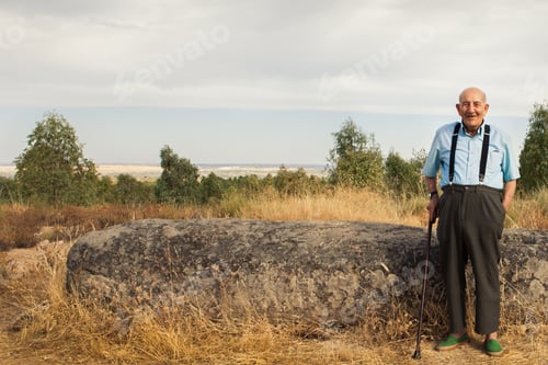 Preview: An elderly man dressed in suspenders leans on a cane next to a menir in the middle of a field.
