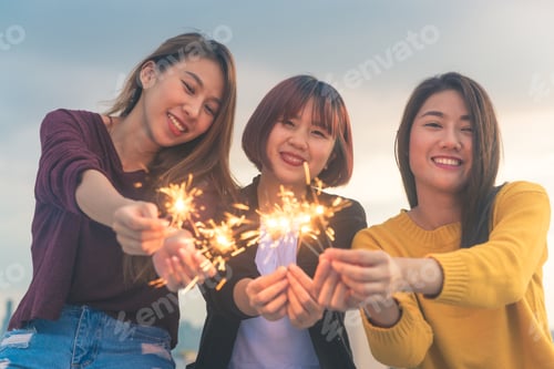 Preview: Happy group of asia girl friends enjoy and play sparkler at roof top party at evening sunset.