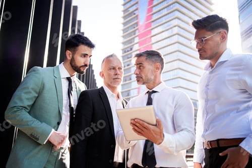 Preview: Group of business men gathered outside offices looking at each other serious consulting on tablet.
