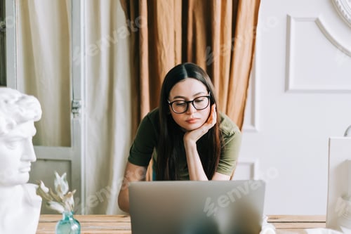 Preview: Young caucasian woman in eyeglasses working on laptop computer in home art studio.