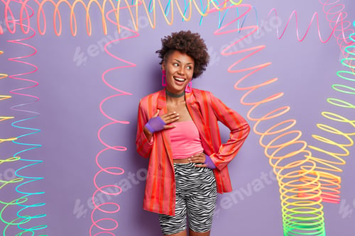 Preview: Positive dark skinned woman laughs happily, keeps hand on hip, dressed in fashionable outfit, poses