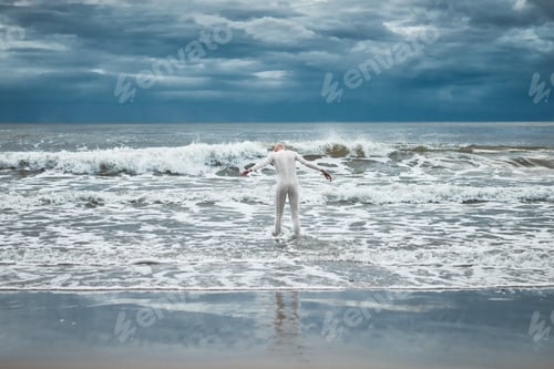 Preview: Hairless girl with alopecia in white futuristic suit goes to cold restless sea on sandy beach
