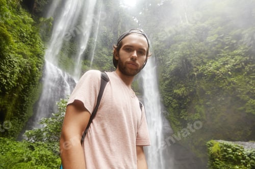 Preview: Stylish handsome hiker in snapback relaxing in the middle of jungle with mountain river in backgroun