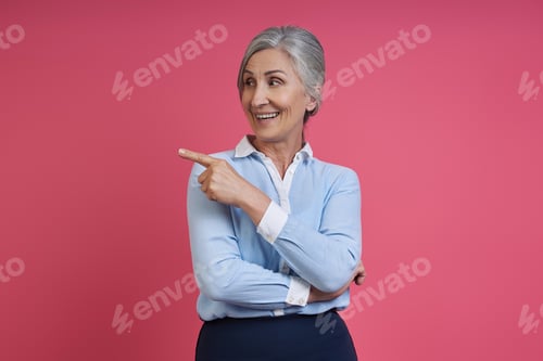 Preview: Confident senior woman pointing away and smiling while standing against pink background