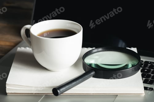 Preview: Cup of coffee, magnifier, notepad and laptop at workplace, close-up.