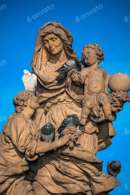 Preview: Statue with mother and child on Charles Bridge in Prague surrounded by pigeons on a clear day