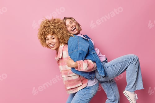 Preview: Horizontal shot of cheerful best women friends give piggyback foolish around dressed in fashionable
